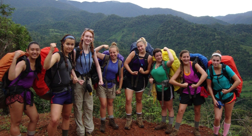 A group of people wearing backpacks pose for a photo in front of green mountains. Many of them are flexing their muscles. 